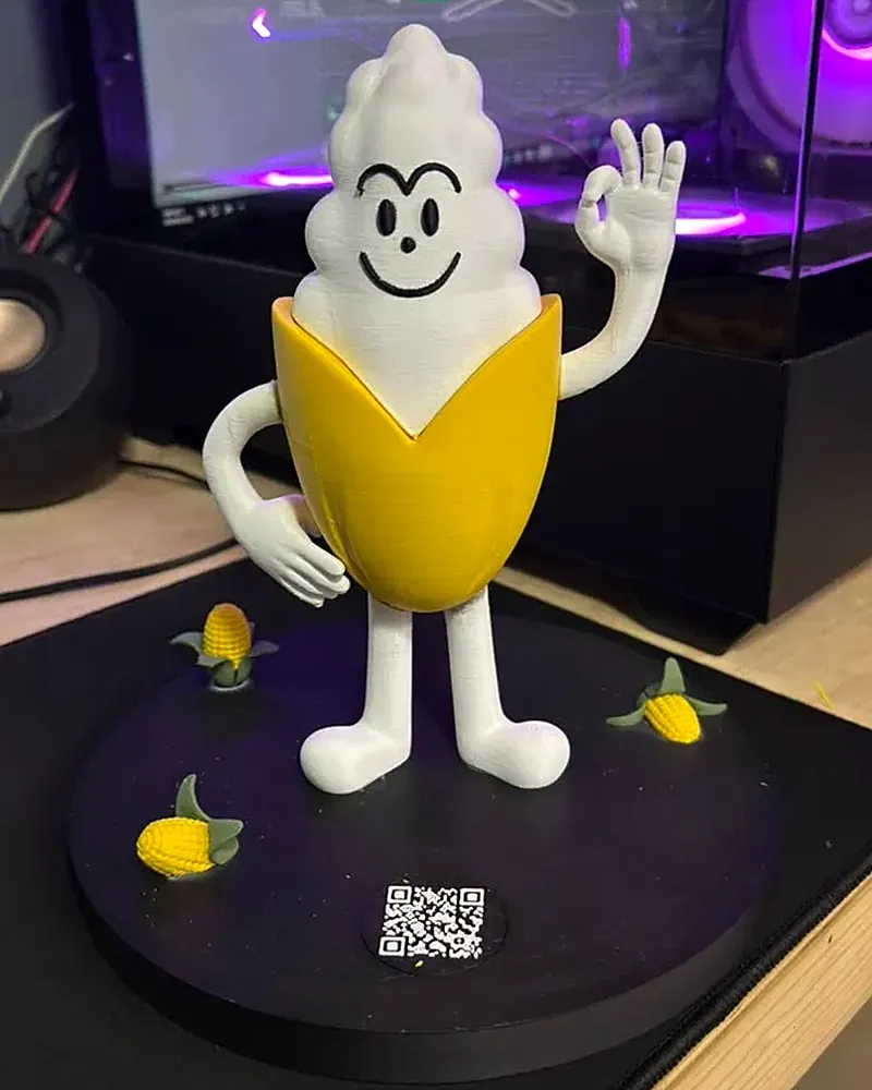 Figura impresa en 3D de Mr. Grano, un elote blanco con hojas amarillas que hace el gesto de OK, sobre una base negra con elotes pequeños y un código QR.